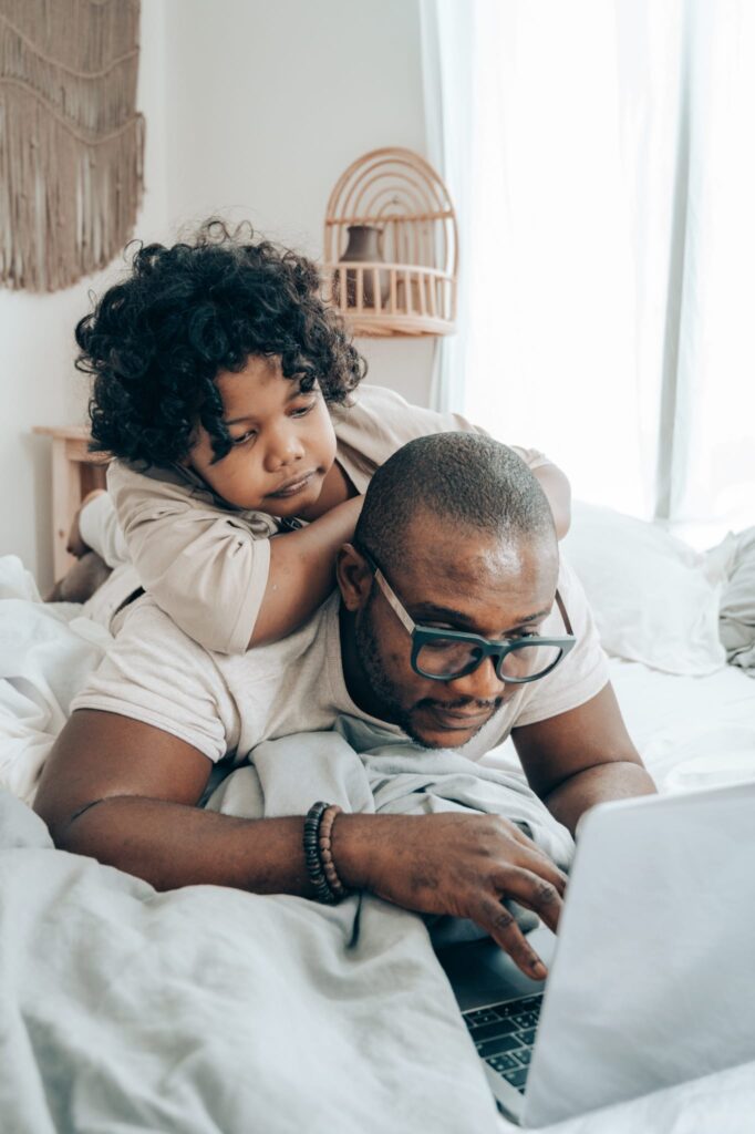 father with child looking over his shoulder while working on laptop in bed